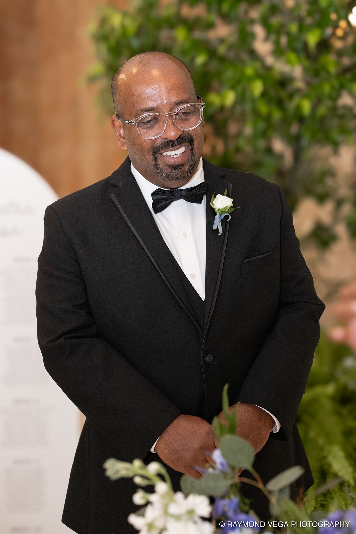 Smiling groom in black tuxedo with bow tie and white boutonniere stands at wedding ceremony altar.