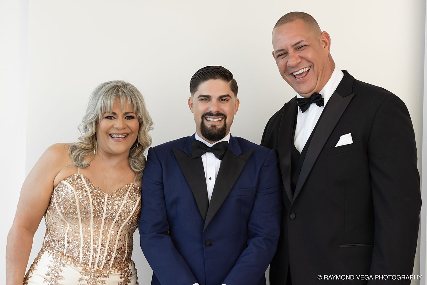 Three elegantly dressed guests smile together at a formal wedding event, wearing black tuxedos and a gold sequin gown.
