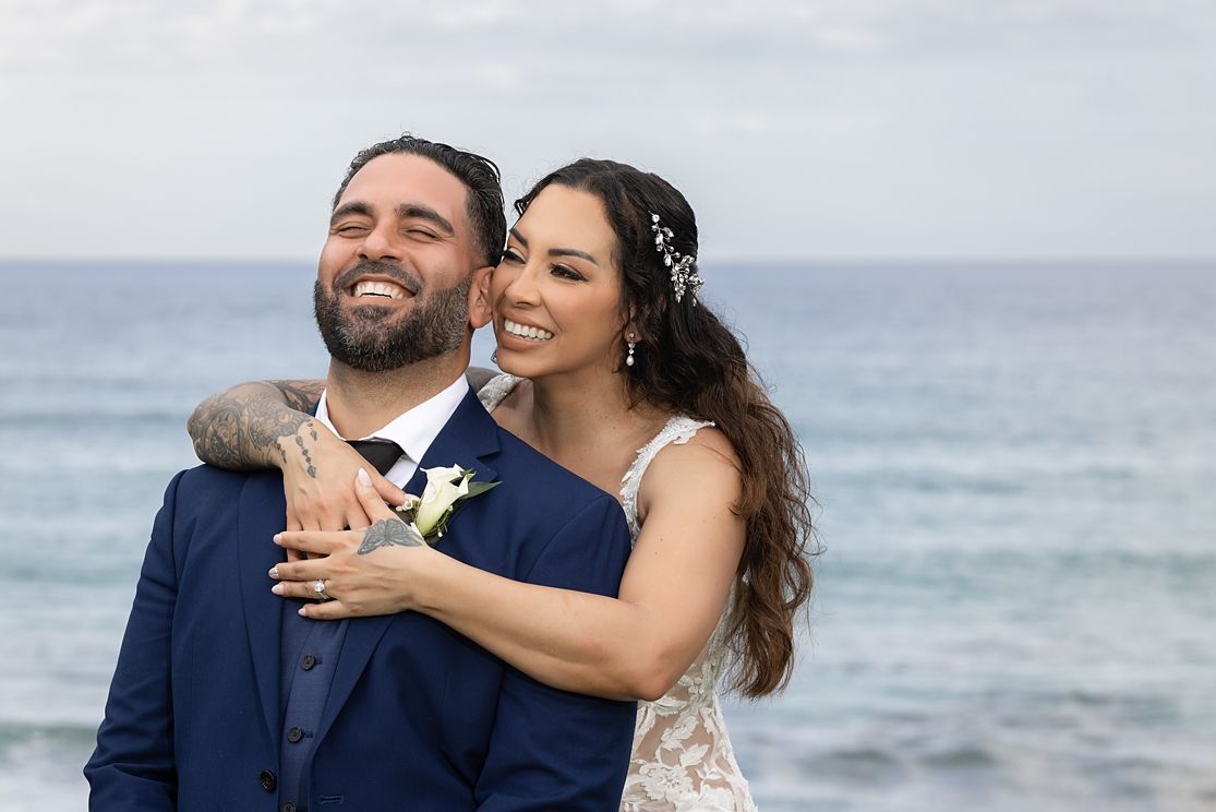 Smiling bride embraces groom in navy suit with ocean backdrop at beach wedding.