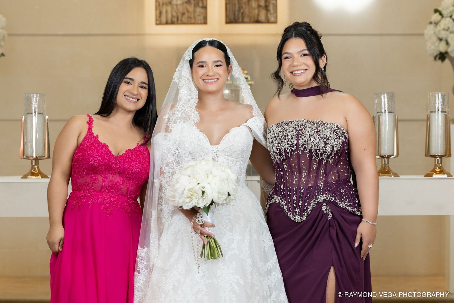 Bride in white lace gown holds bouquet flanked by bridesmaids in pink and purple dresses at church altar.