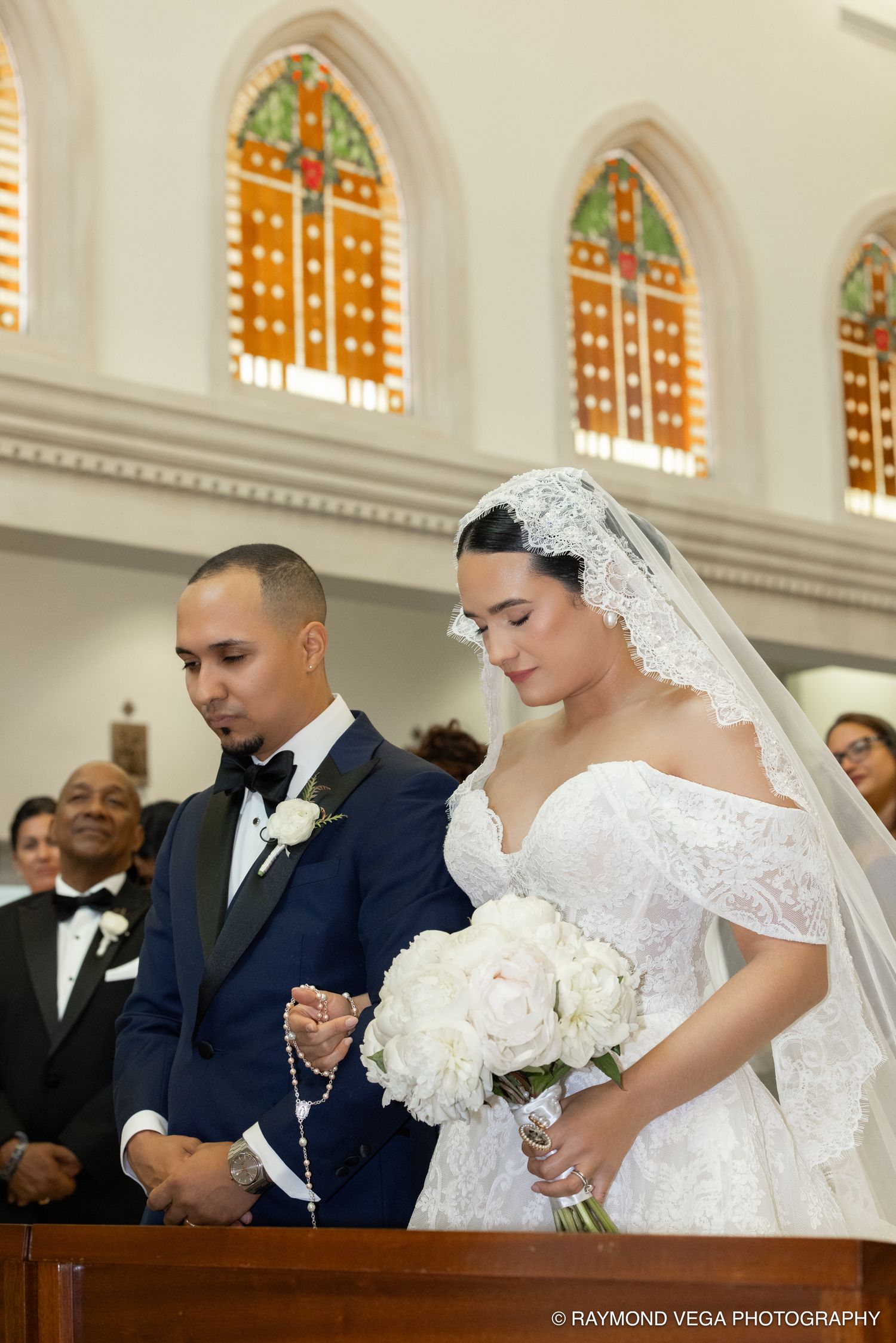 Bride and groom praying at church altar, bride in lace gown with veil holding white peonies bouquet, groom in navy tuxedo.