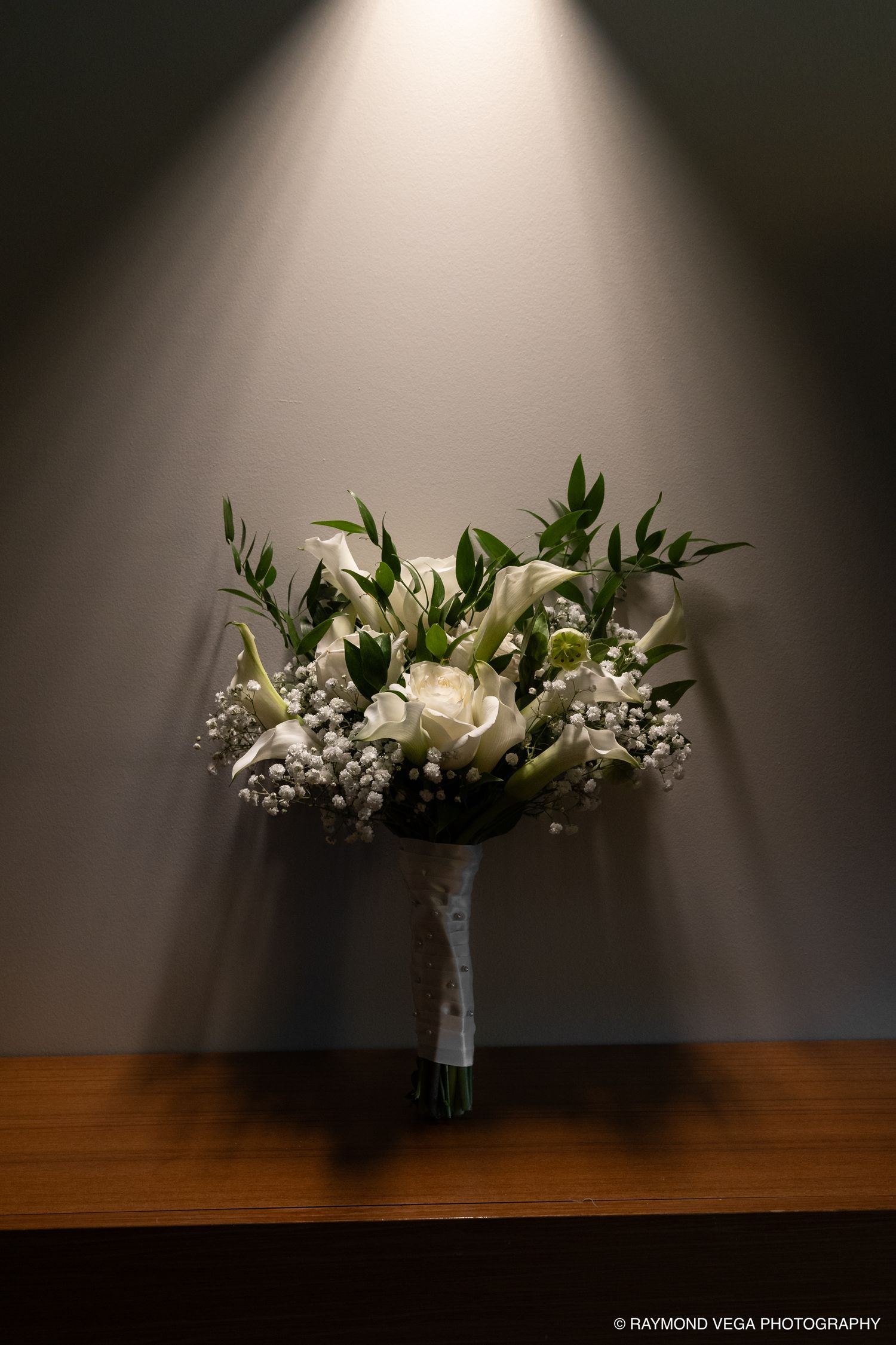 White wedding bouquet with roses, calla lilies, and greenery displayed on wooden surface under spotlight against gray wall.