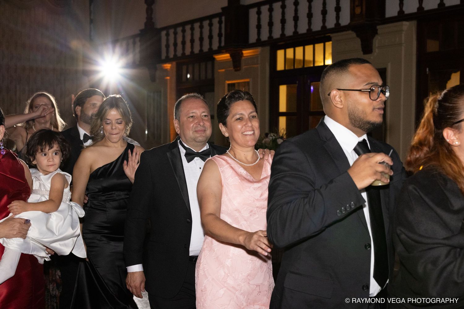 Elegantly dressed guests smile and pose at a formal evening event, with a man in a tuxedo and woman in pink standing prominently.