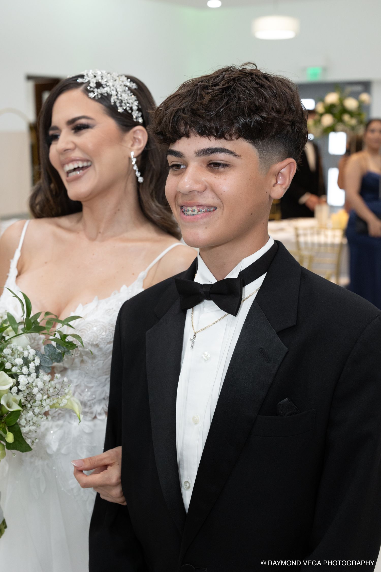 A young boy in a tuxedo smiles beside a laughing bride in a white gown holding a bouquet at a wedding reception.