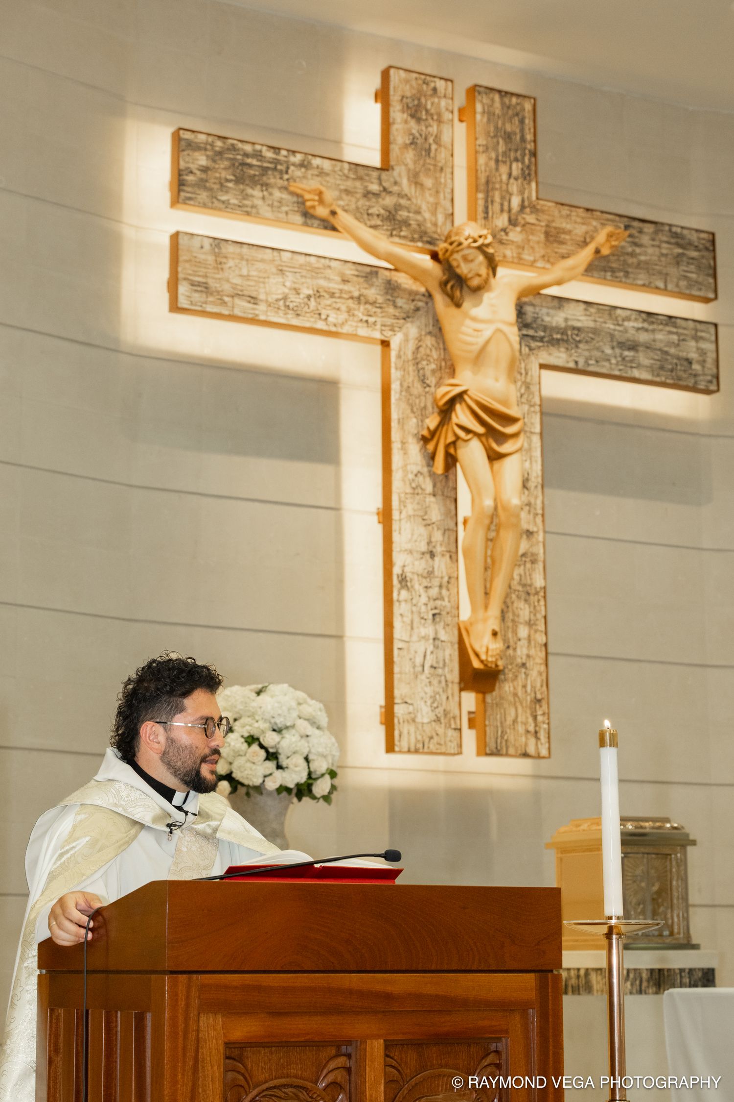 Priest reads at wooden pulpit before large crucifix with Jesus figure in a Catholic church.