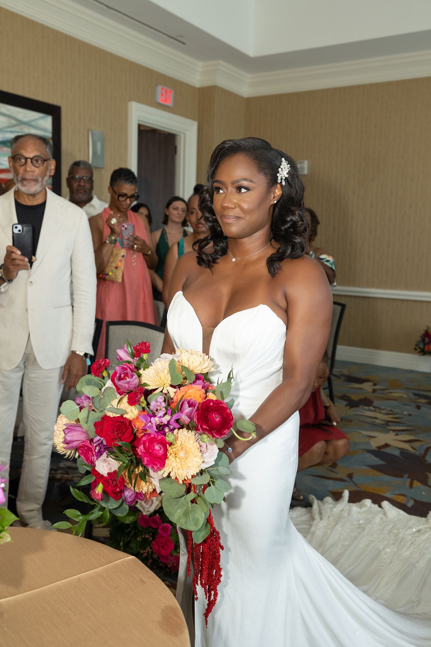 Bride in white strapless gown holds vibrant bouquet of red, pink, and orange flowers during indoor wedding ceremony.