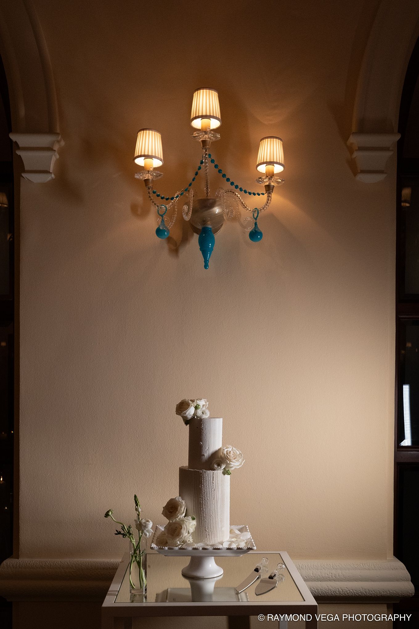 Elegant white tiered wedding cake displayed on a mirrored table beneath a decorative wall sconce with blue accents.