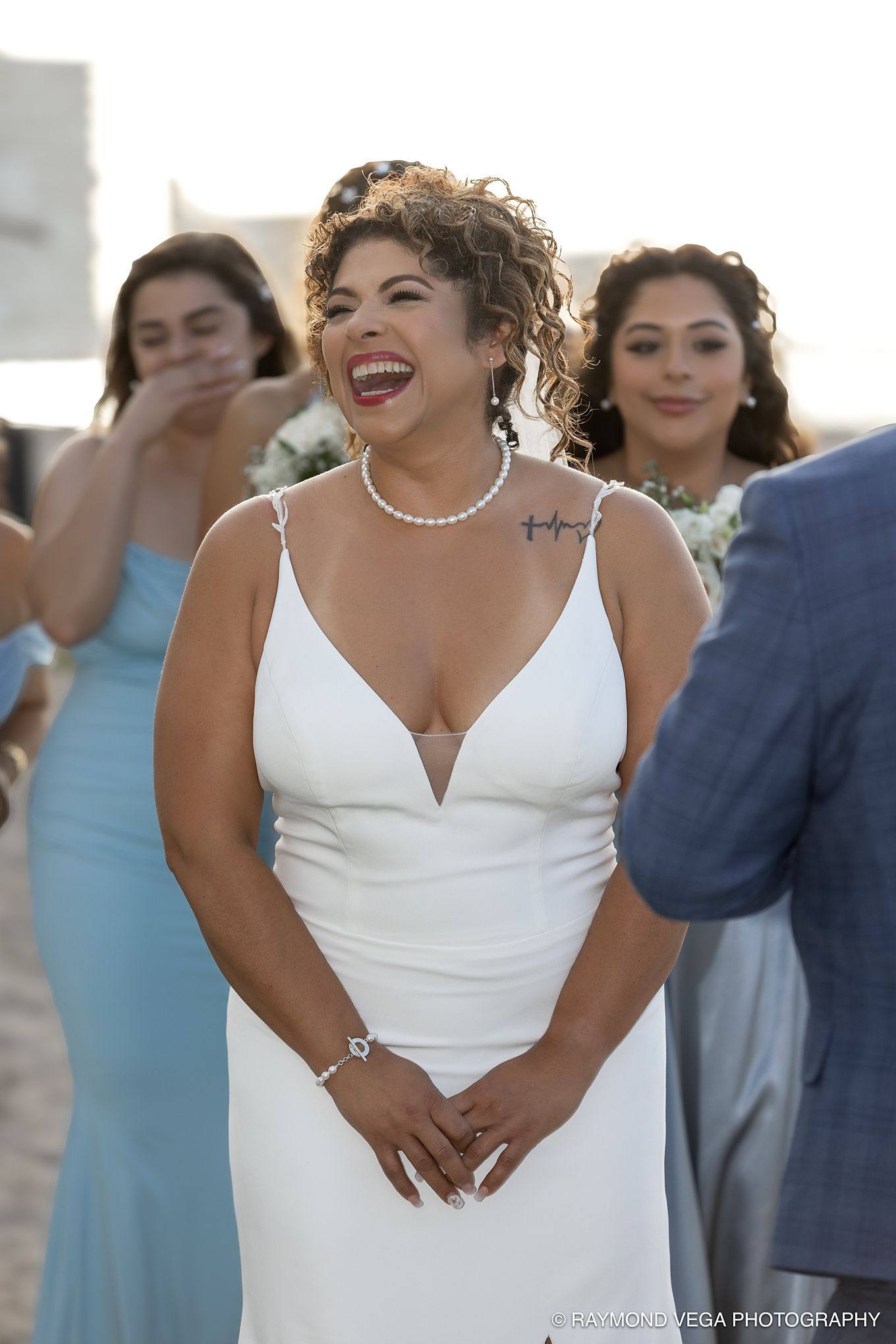 Bride in white gown laughing joyfully at outdoor wedding ceremony with bridesmaids in blue dresses behind her.