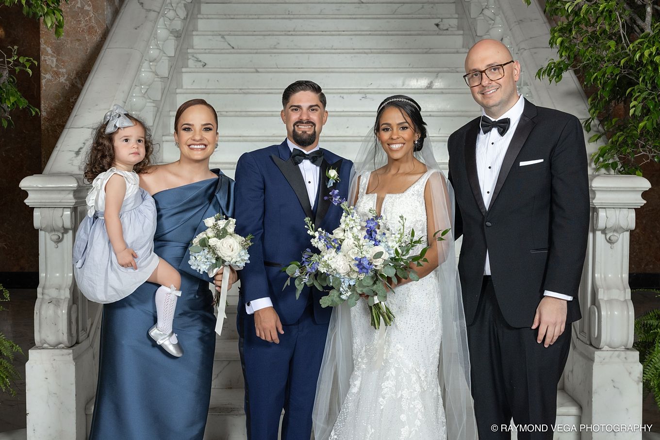 Wedding party poses on elegant stone steps, bride in white lace gown holding blue bouquet with groom and guests.