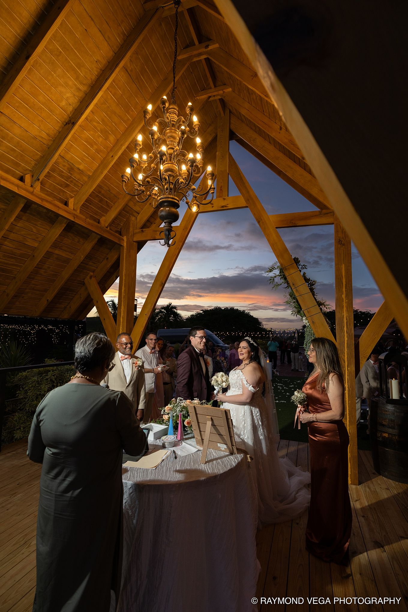 Wedding ceremony under wooden A-frame pavilion with chandelier at sunset, bride and groom at altar with officiant.
