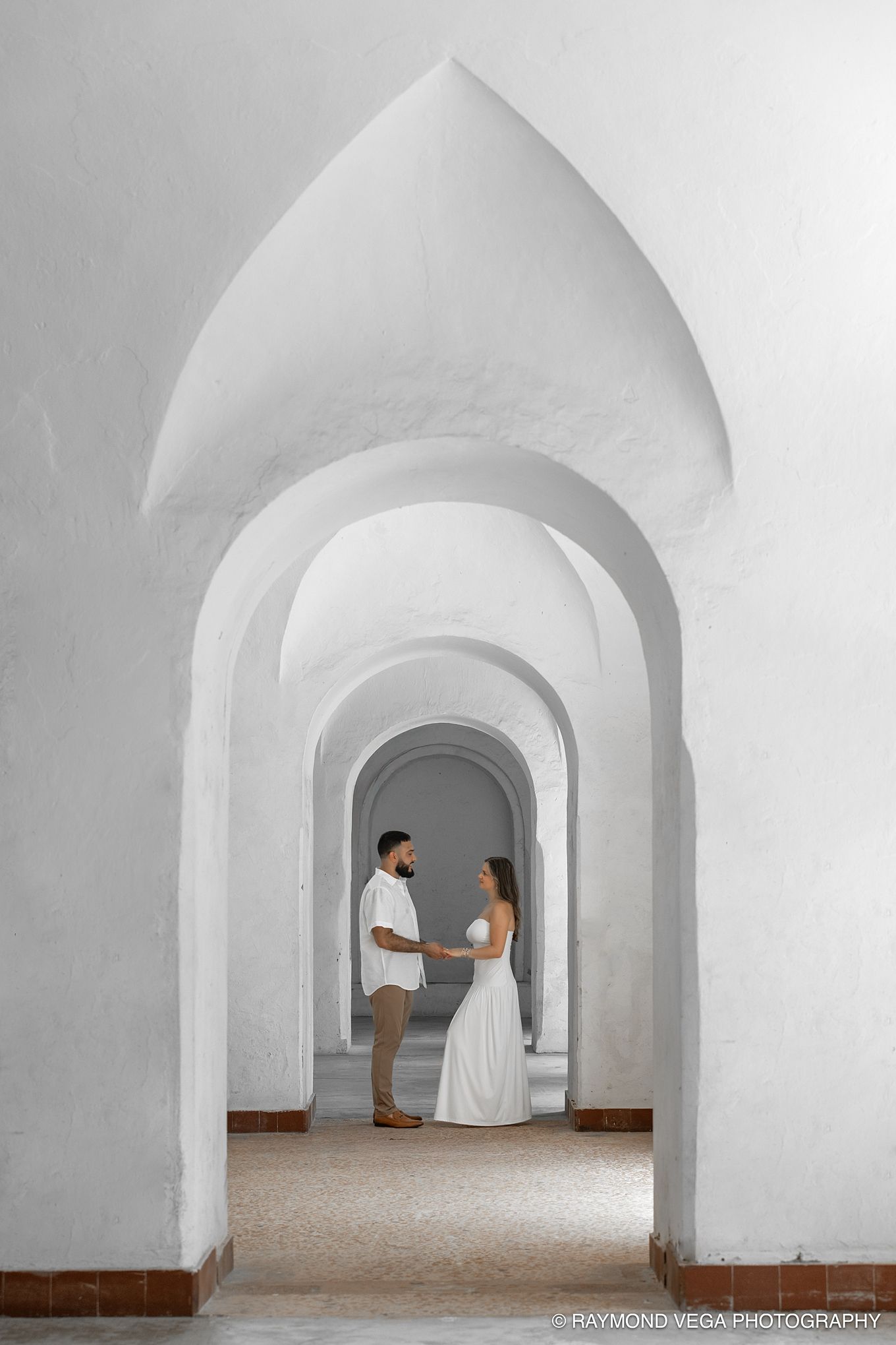 Couple in white attire holding hands under elegant arched corridor, captured by Raymond Vega Photography.