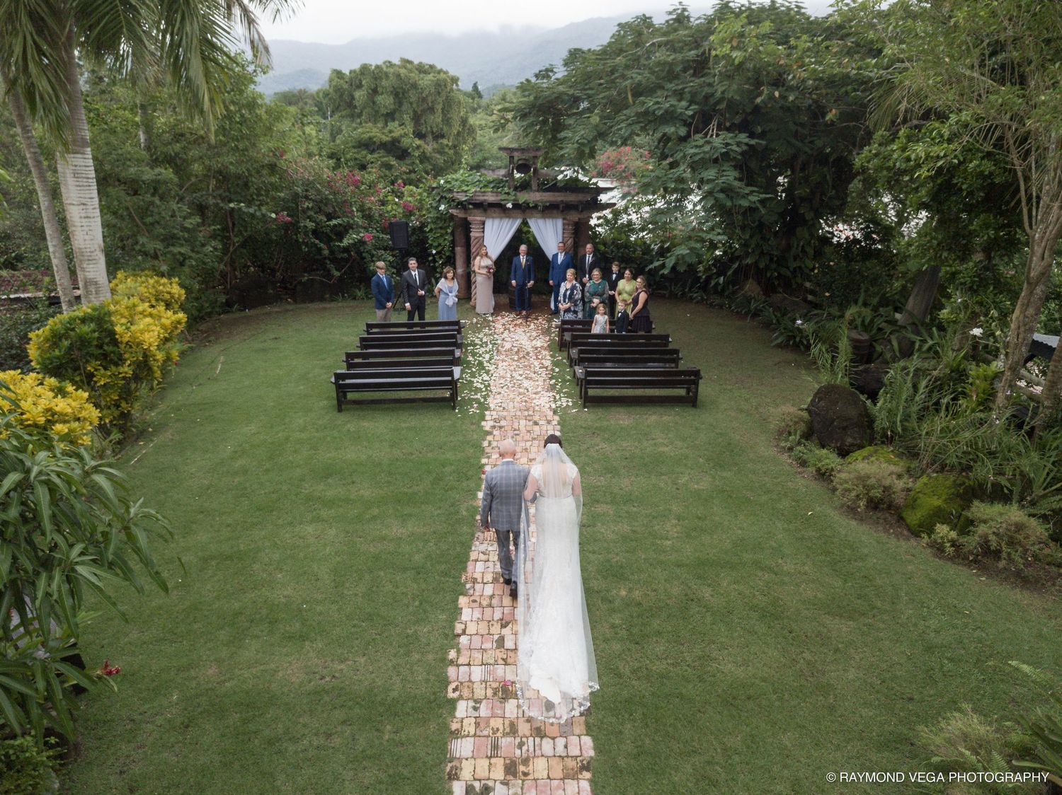 Aerial drone view of elegant garden wedding ceremony with lush greenery and stone-lined aisle.