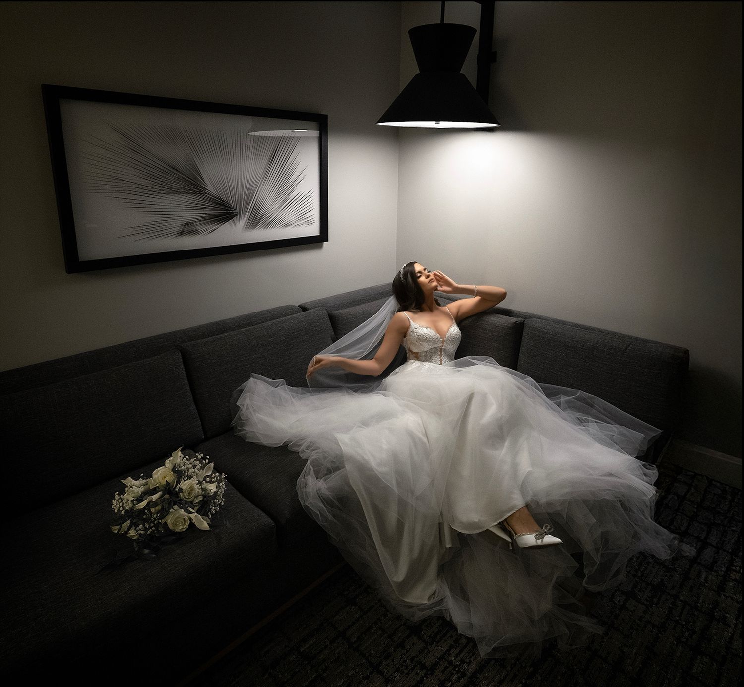 Bride in white wedding gown relaxing on gray sofa under pendant light, bouquet nearby, dramatic dark room ambiance.
