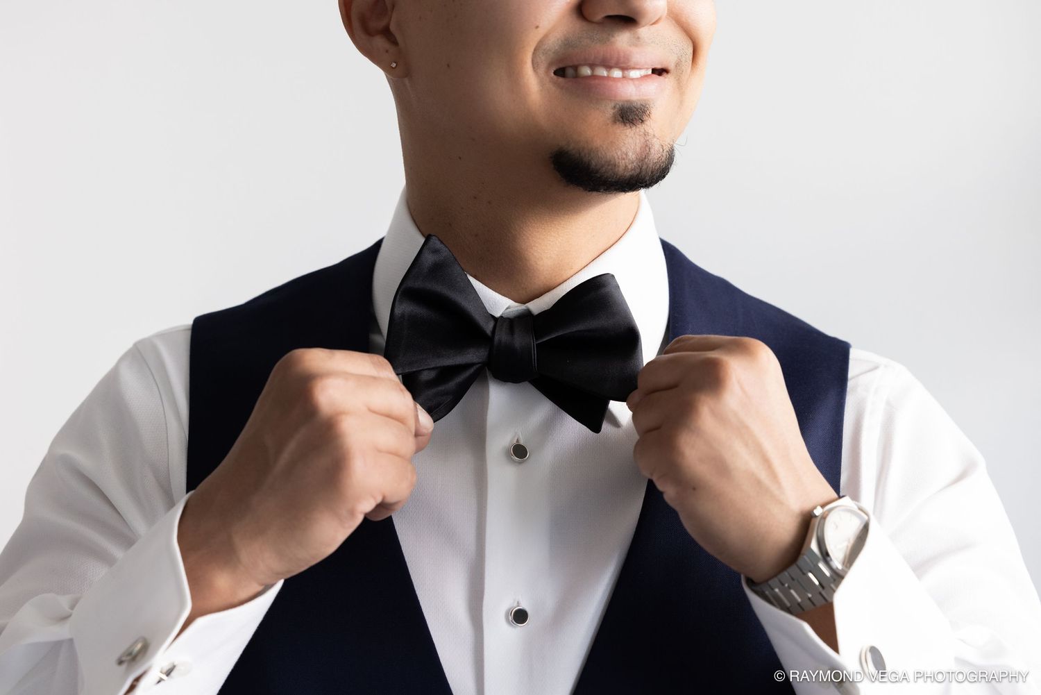 Smiling man in navy vest and white shirt adjusting black bow tie, wearing a silver watch, against gray background.