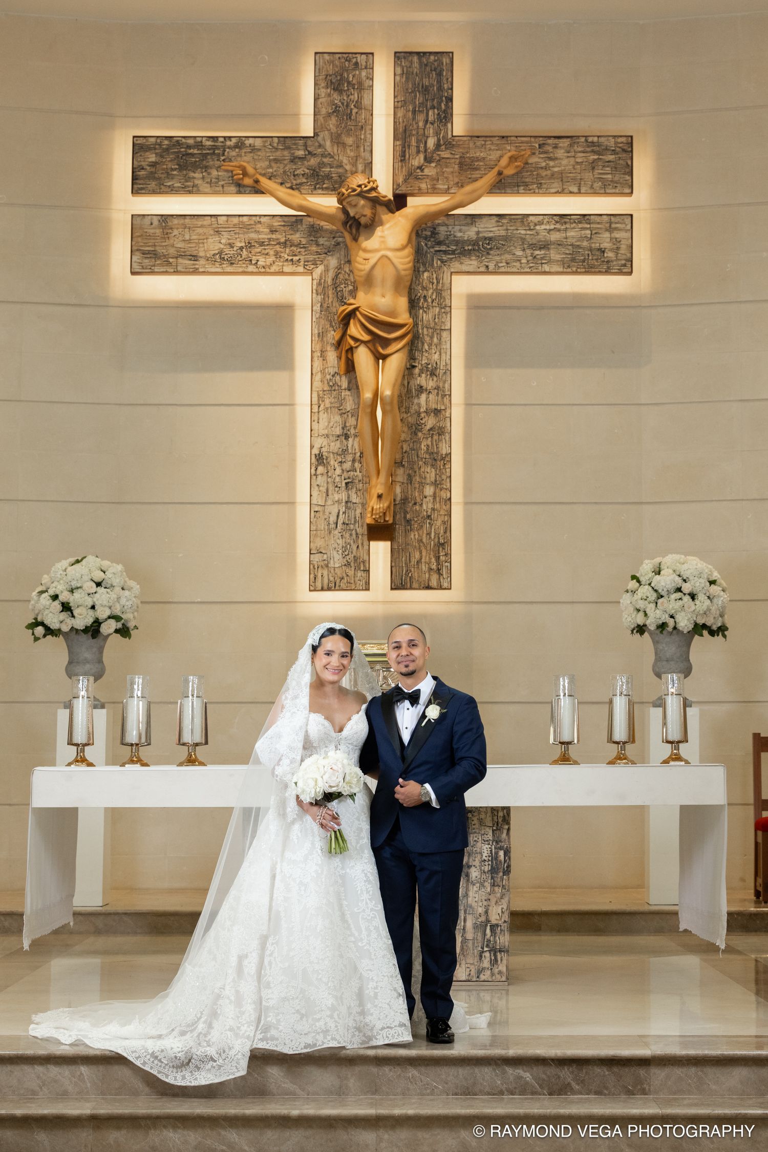 Bride and groom posing at church altar beneath crucifix with white floral arrangements.