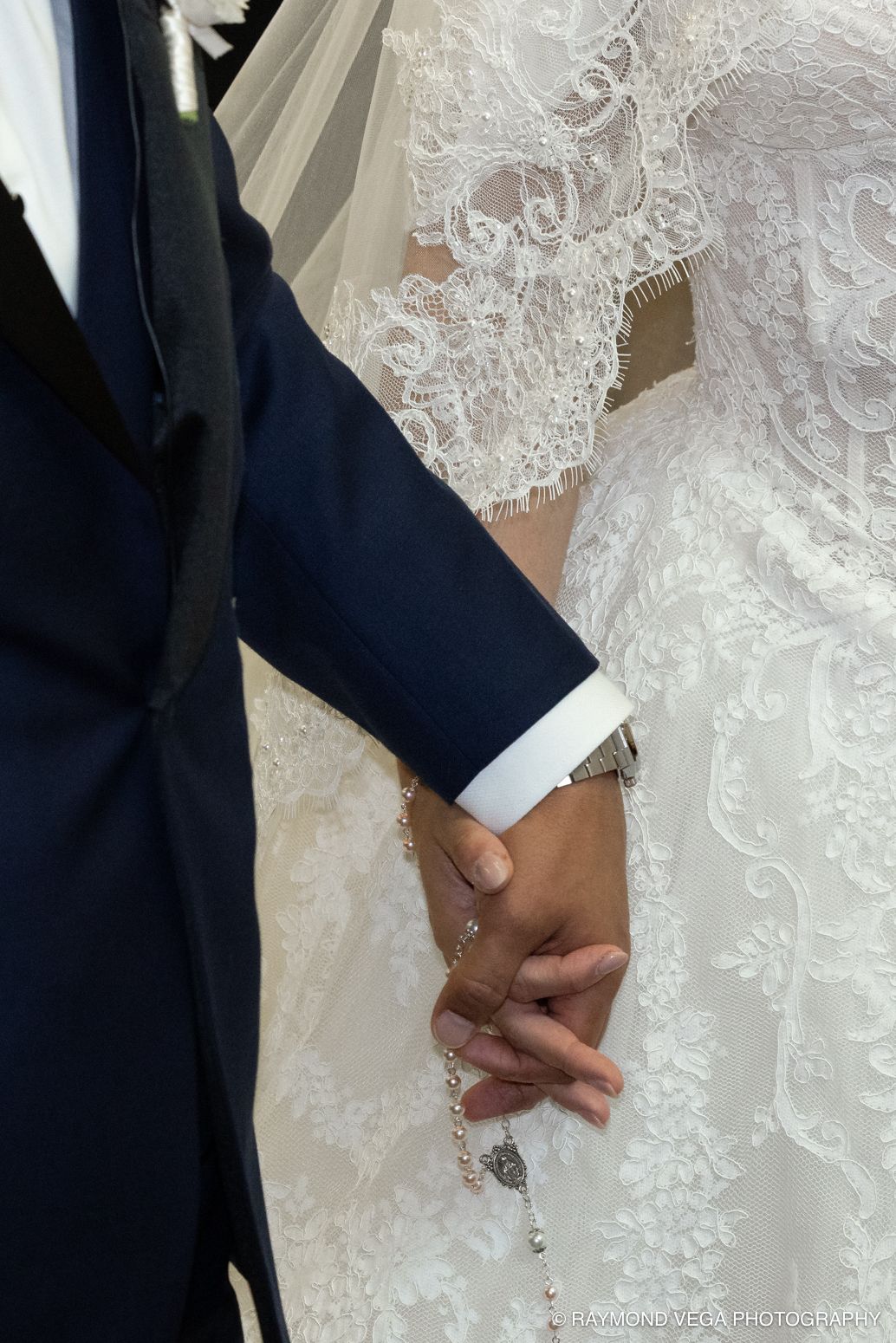 Bride and groom holding hands, showcasing lace wedding dress, blue suit, and pearl rosary bracelet during wedding ceremony.