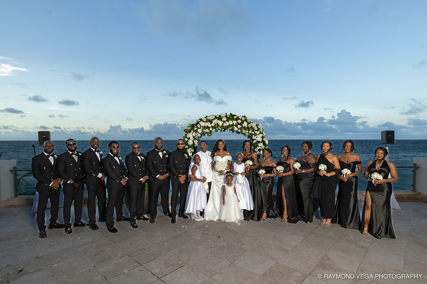 Wedding party poses outdoors by the ocean at dusk, with bride, groom, groomsmen in tuxedos, and bridesmaids in black gowns.