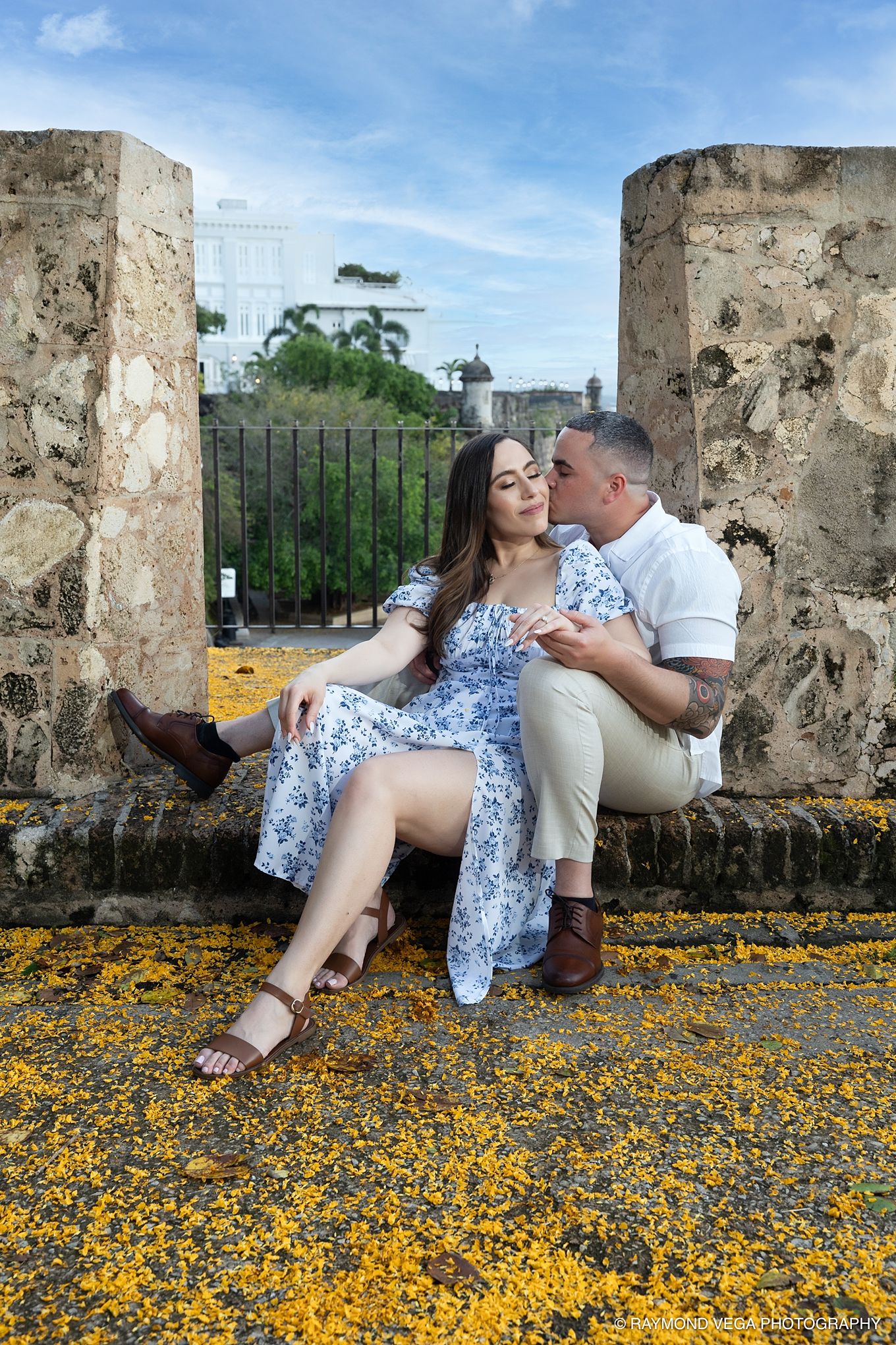 Couple sharing a romantic kiss on stone steps surrounded by yellow flowers at a historic outdoor location. - Love Story