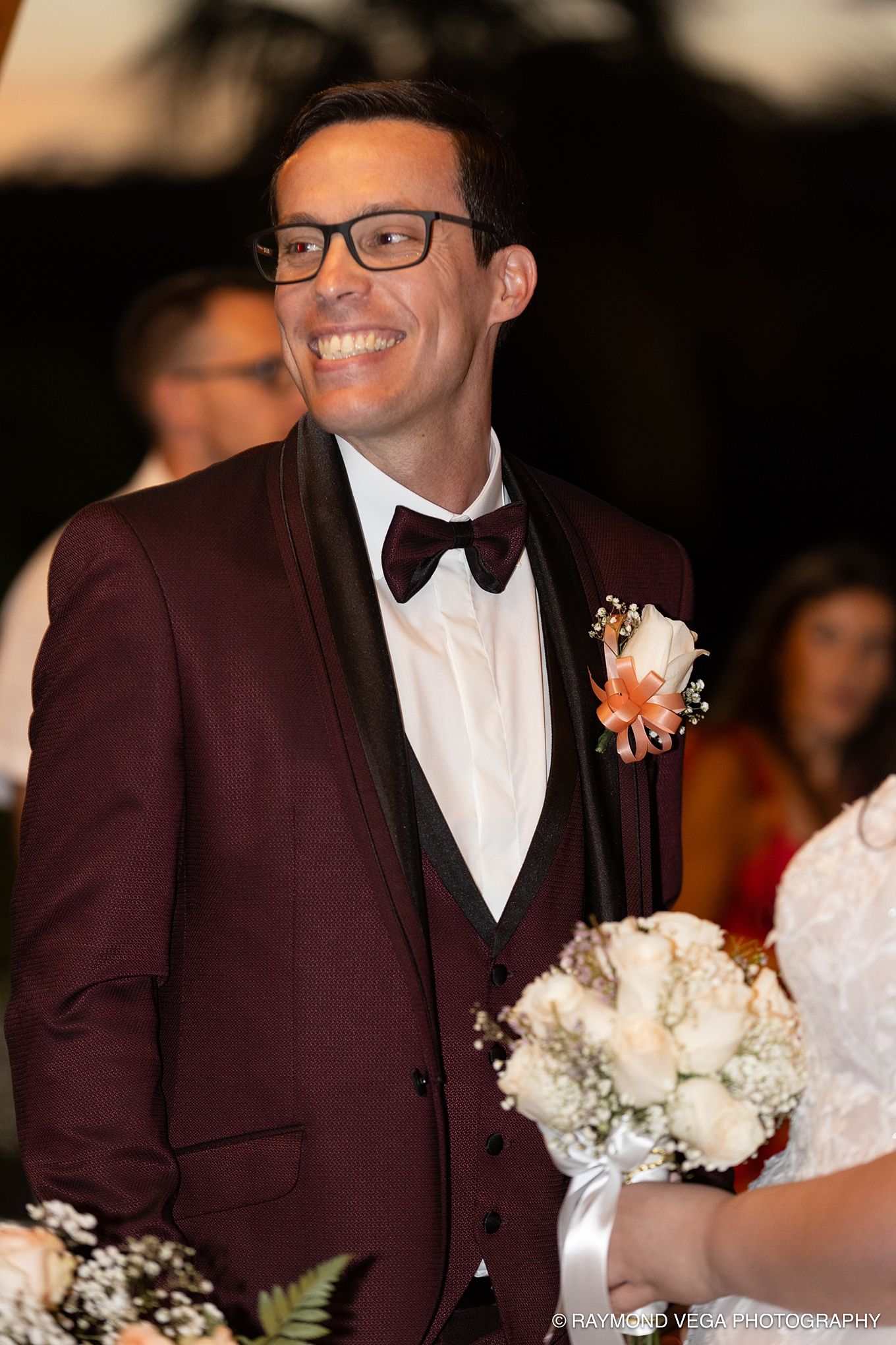 Smiling groom in burgundy tuxedo with bow tie and boutonniere at wedding ceremony.