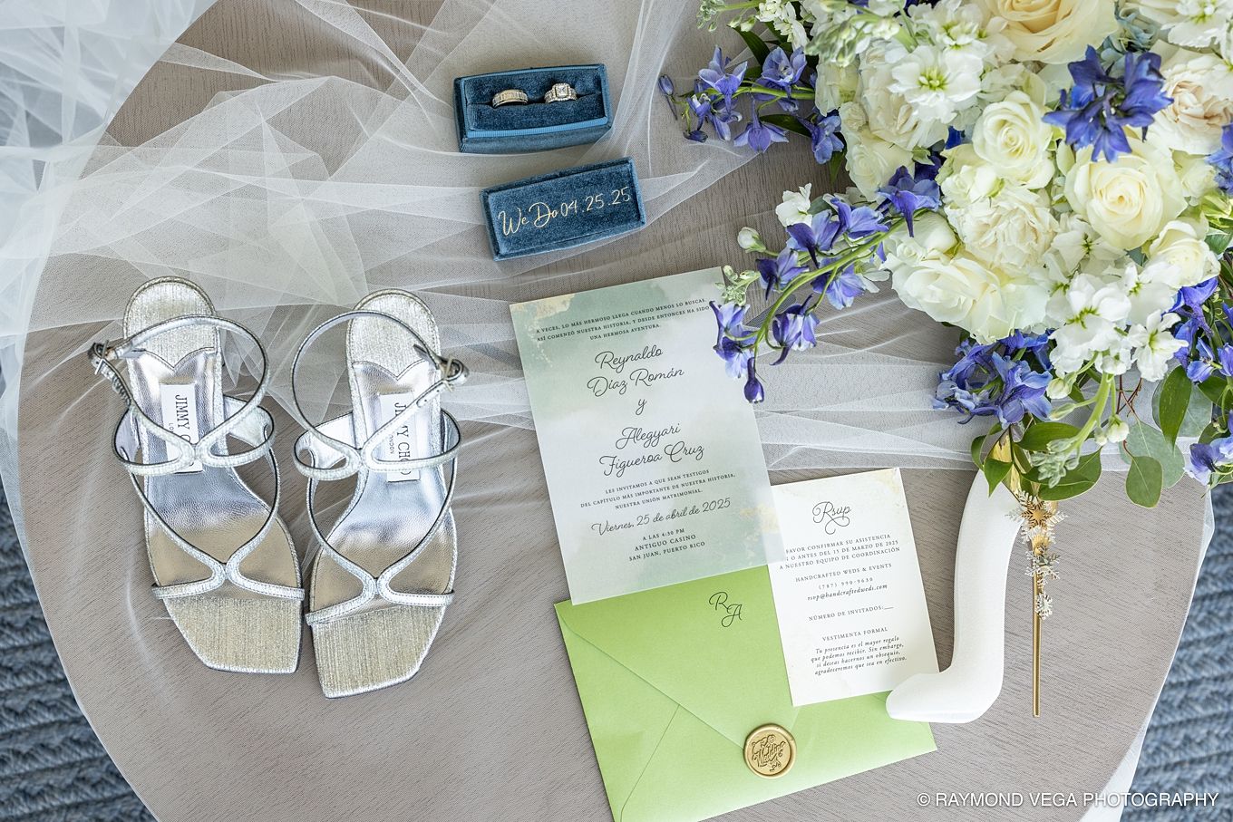 Wedding flat lay featuring silver sandals, green invitations, blue floral bouquet, and ring boxes on a neutral surface. Hotel la Concha