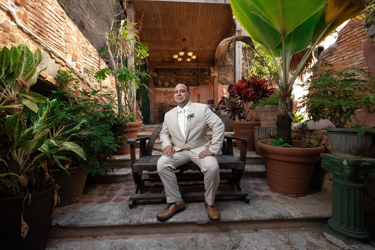 Man in white suit sitting on wooden bench in lush courtyard with brick walls and tropical plants.