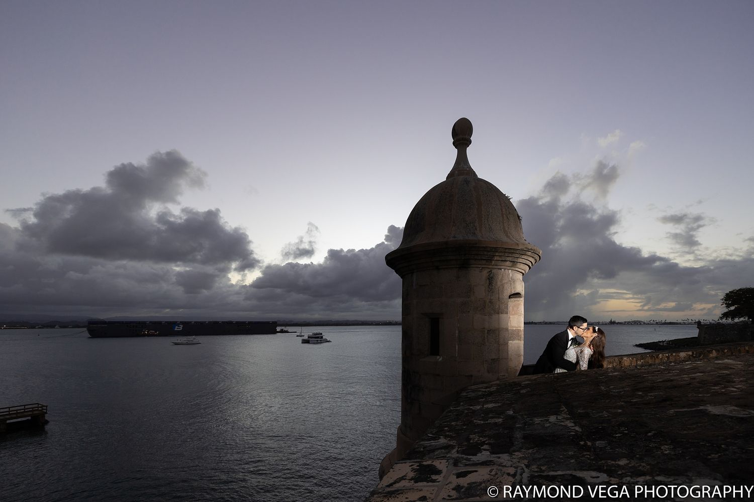 Couple sharing a romantic kiss beside a historic stone turret overlooking a serene bay at dusk in San Juan.