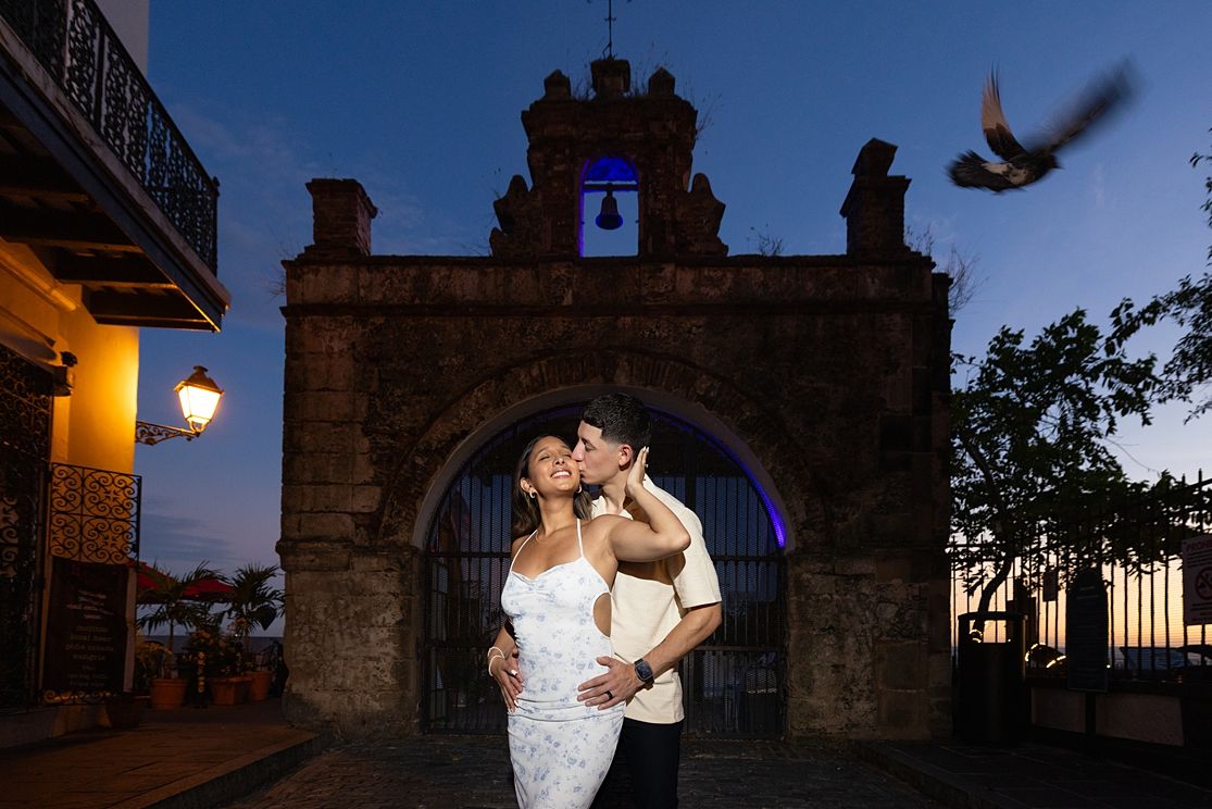 Couple sharing a romantic kiss at dusk before a historic stone gate in Old San Juan, Puerto Rico.