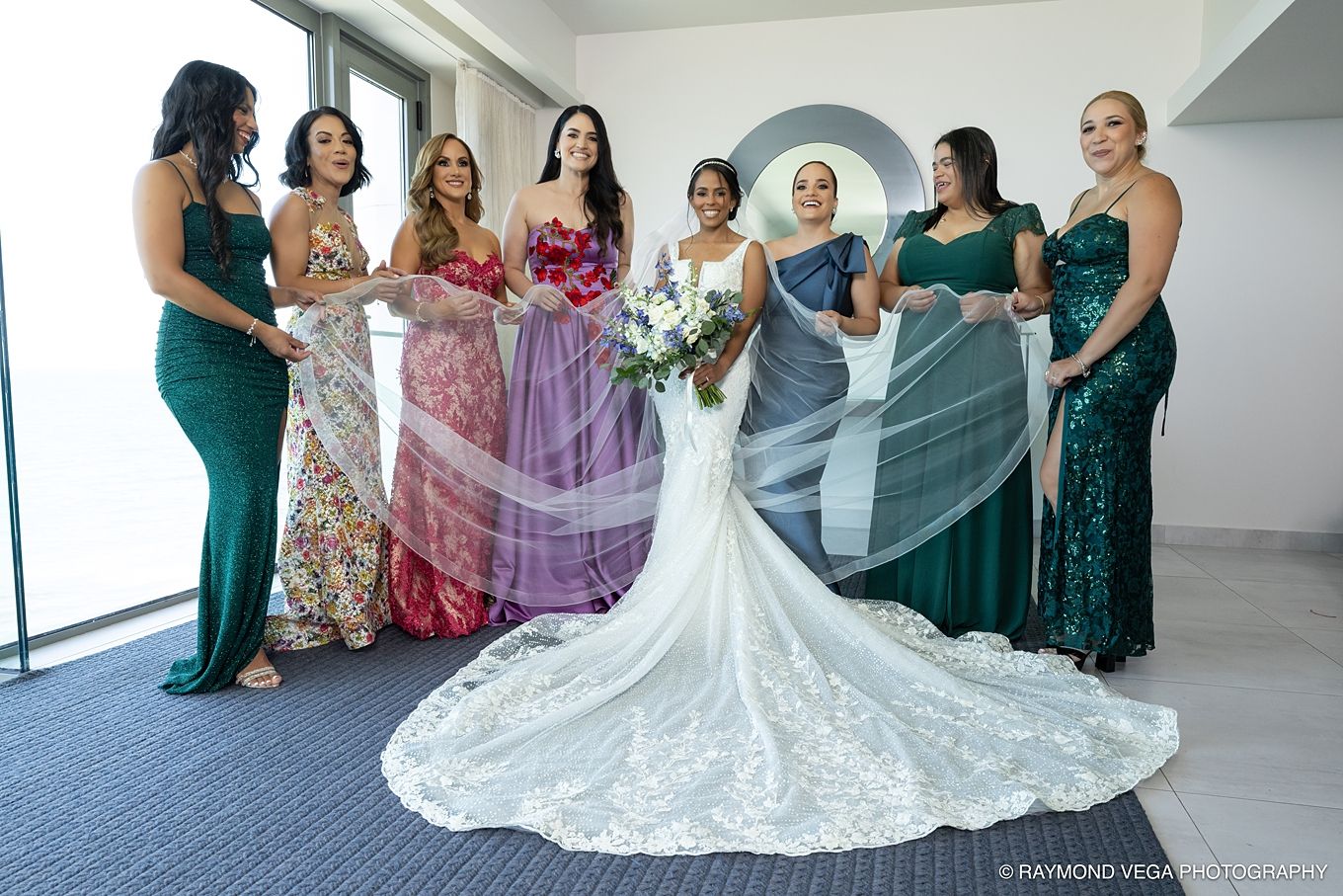 Bride in white lace gown poses with six bridesmaids in green and colorful dresses, holding a bouquet indoors. Hotel la Concha Wedding