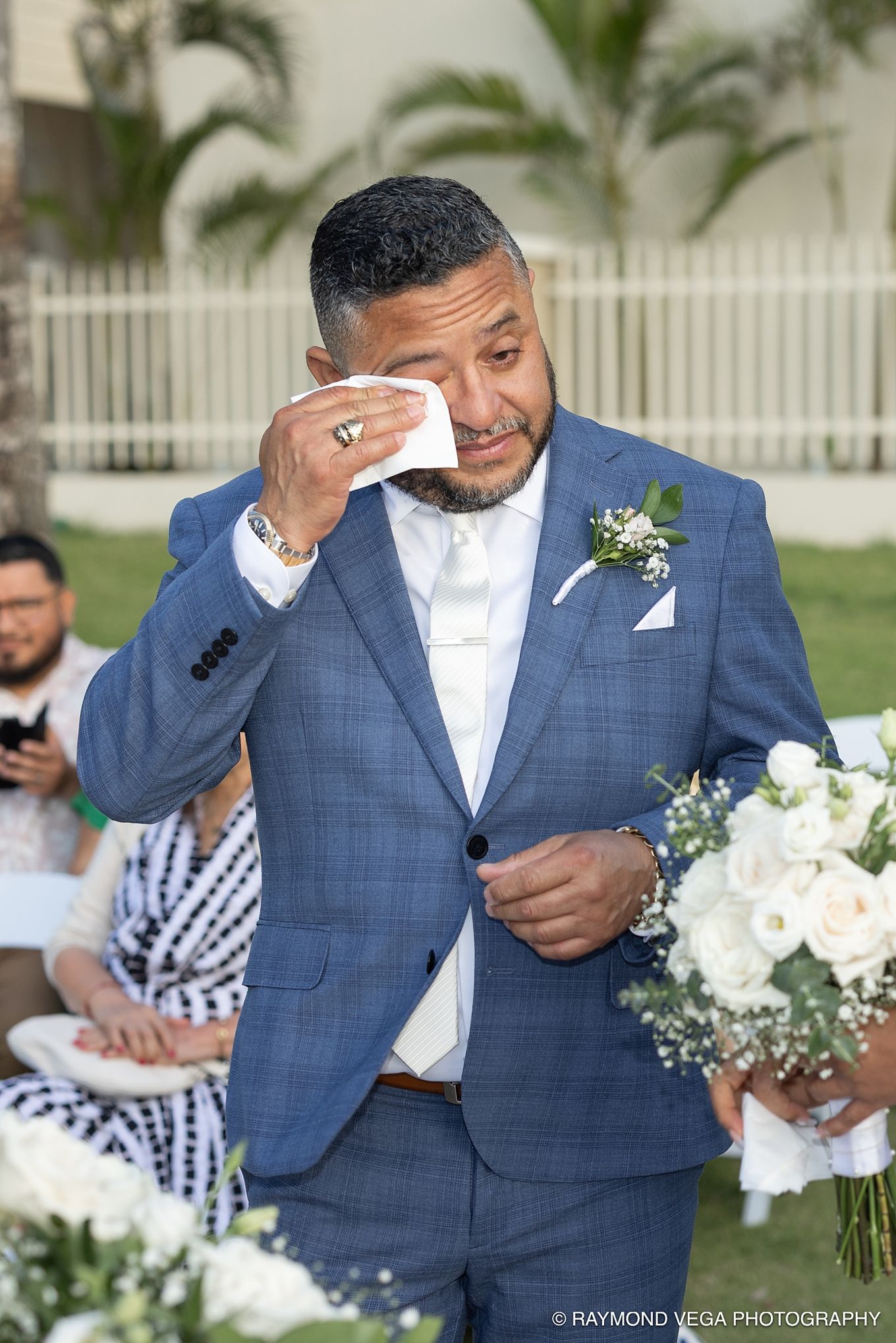 Emotional groom in blue suit wiping tears holding white roses bouquet during outdoor wedding ceremony.