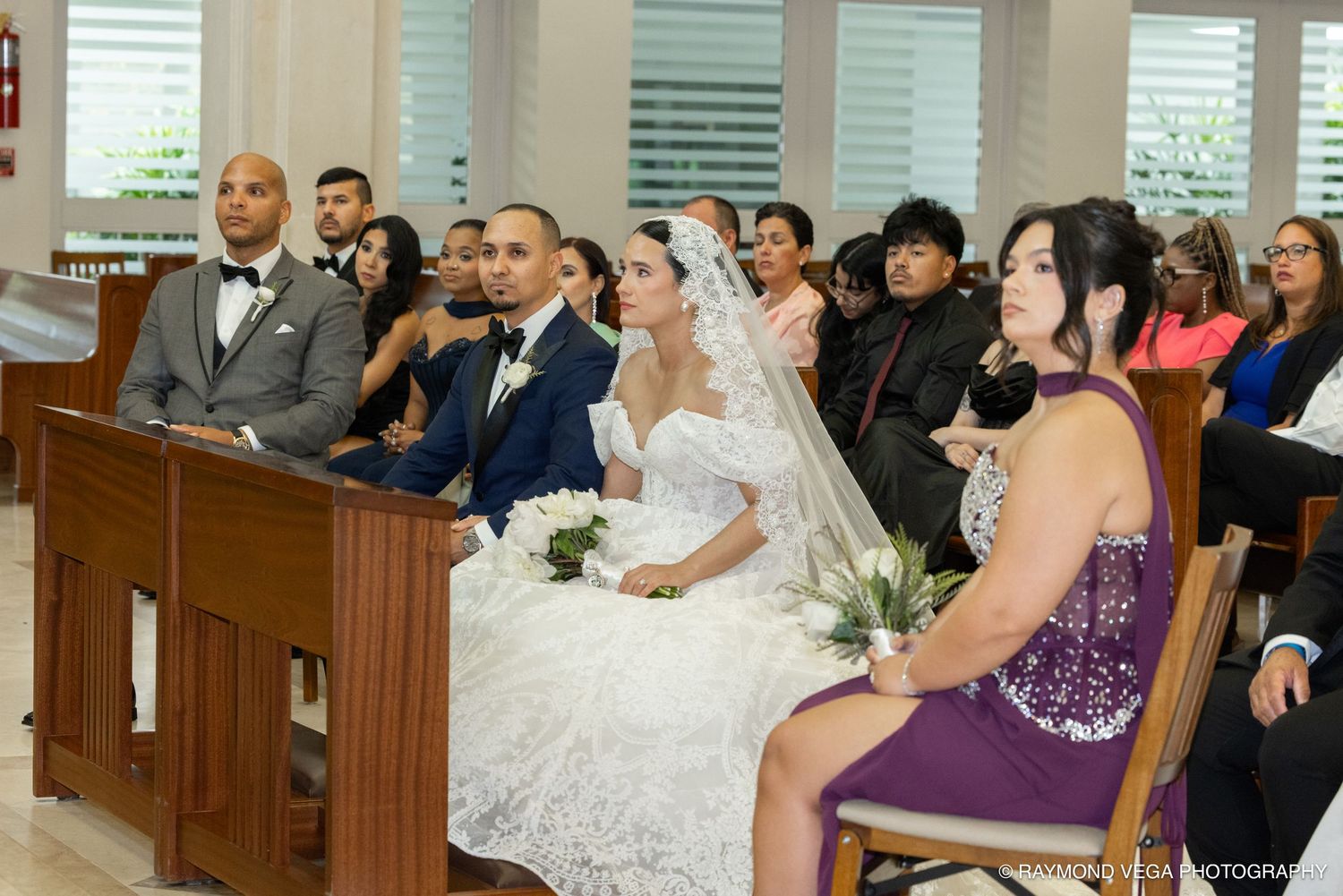Bride and groom seated at church altar during wedding ceremony with guests in pews and bridesmaid in purple dress.