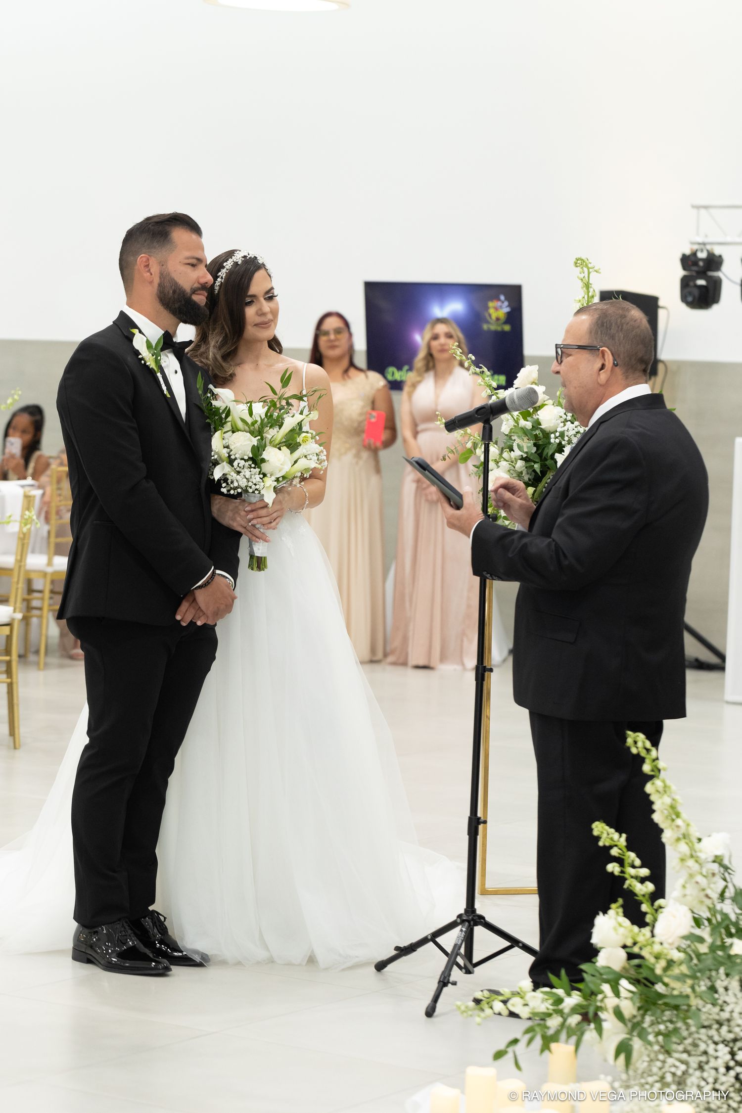Couple exchanges wedding vows at altar with officiant and bridesmaids in elegant white venue.
