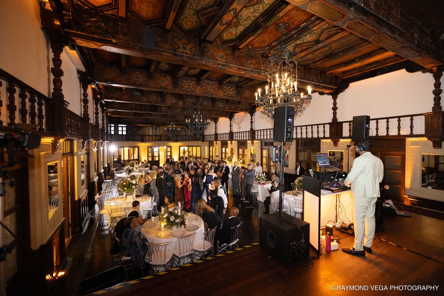 Elegant wedding reception in a grand historic hall with chandeliers, guests mingling, and a DJ performing at the venue.