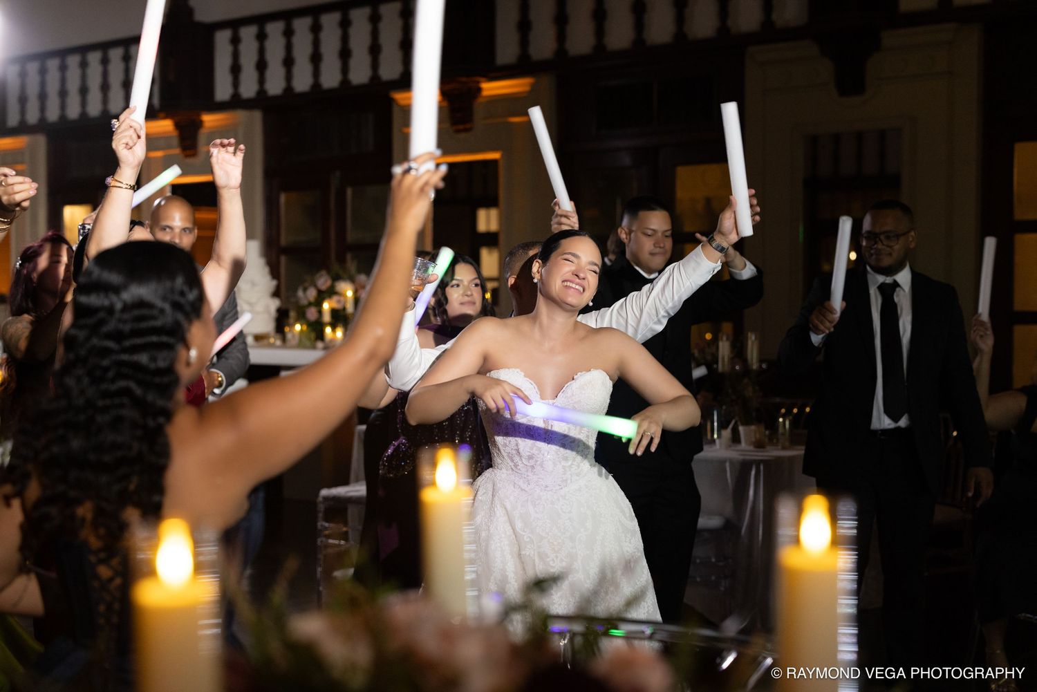 Bride and groom celebrate at wedding reception with guests raising glow sticks, candlelit tables in elegant venue.