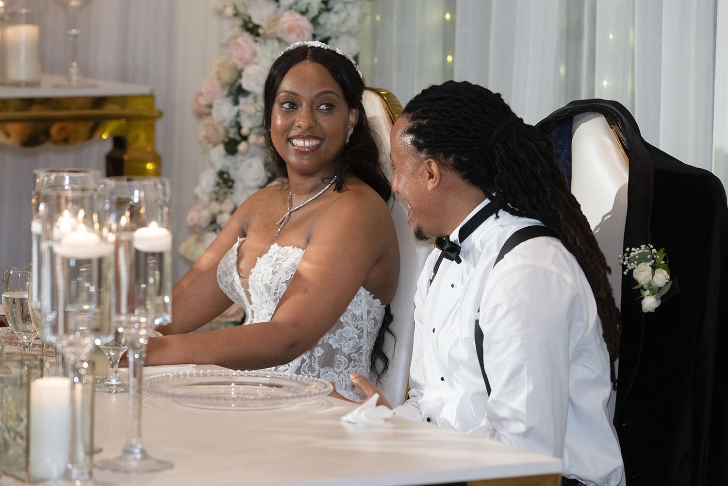 Bride and groom seated at wedding reception table with candles and floral decor, smiling joyfully.