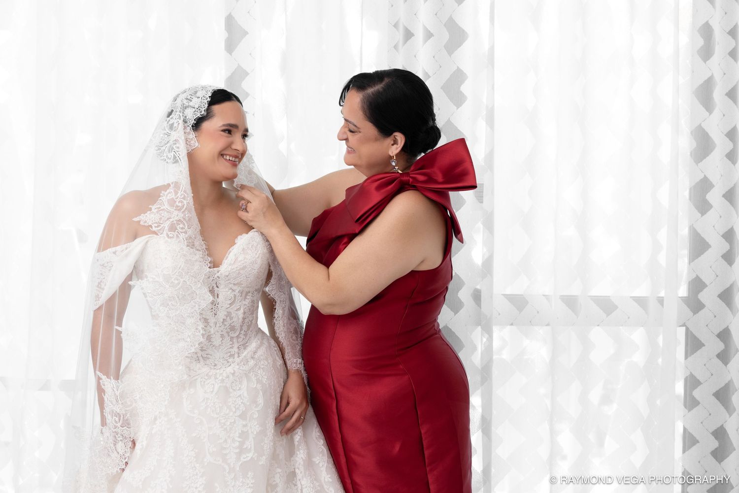 Bride in white lace gown and veil being helped by woman in red dress near sheer curtains.