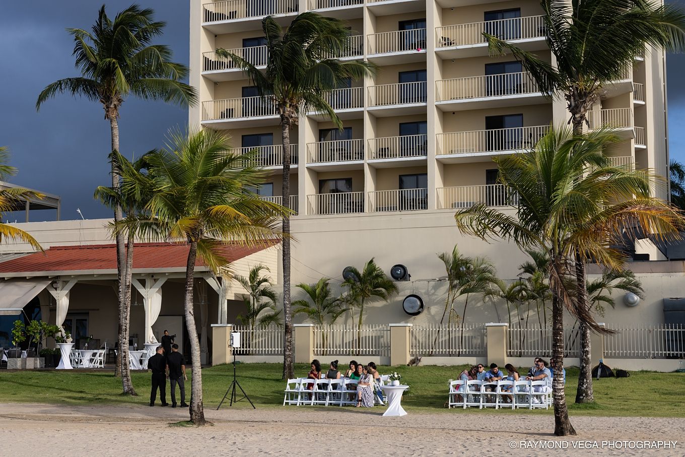 Outdoor beach wedding reception setup with white chairs near a tropical hotel surrounded by palm trees.