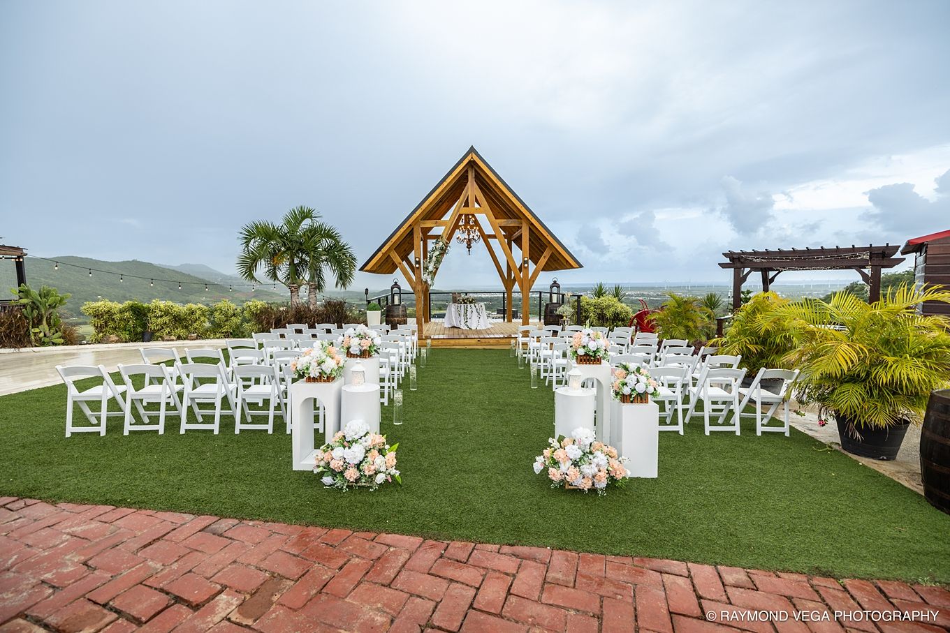 Outdoor wedding ceremony setup with white chairs, floral arrangements, and a wooden gazebo overlooking lush green hills.