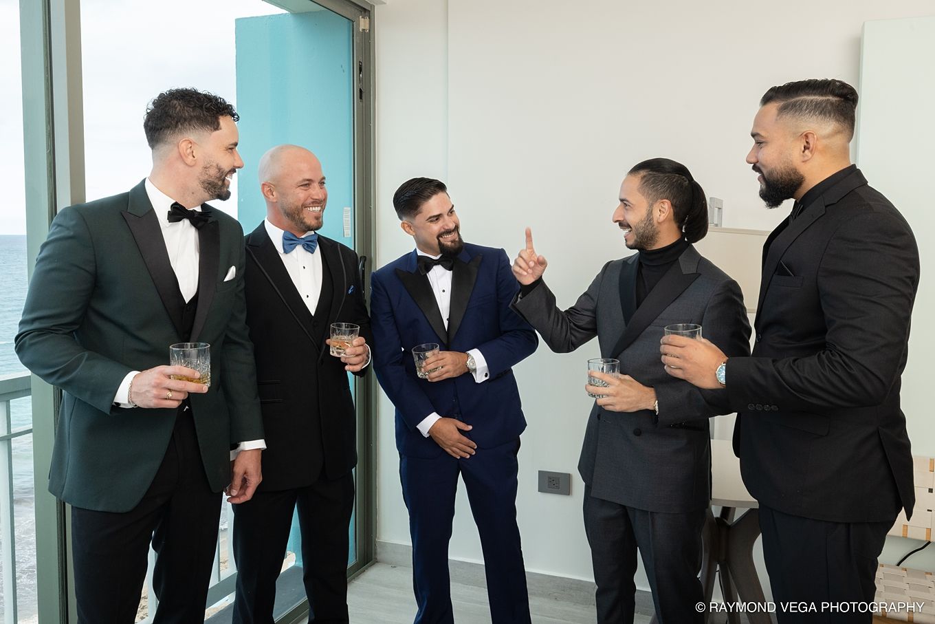 Five men in tuxedos and suits laughing and toasting drinks at a formal wedding celebration. Hotel la Concha Wedding