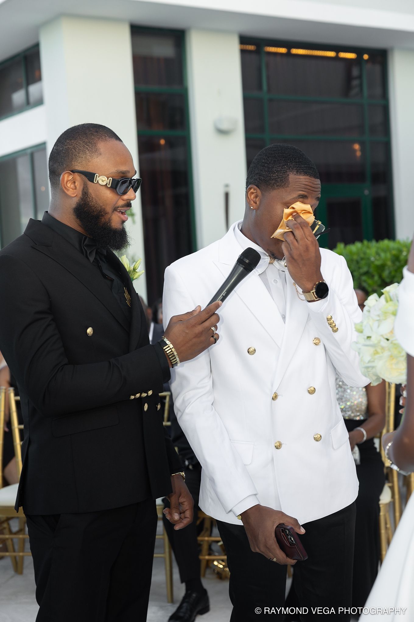 Groom in white blazer wipes tears at outdoor wedding ceremony while man in black suit holds microphone.