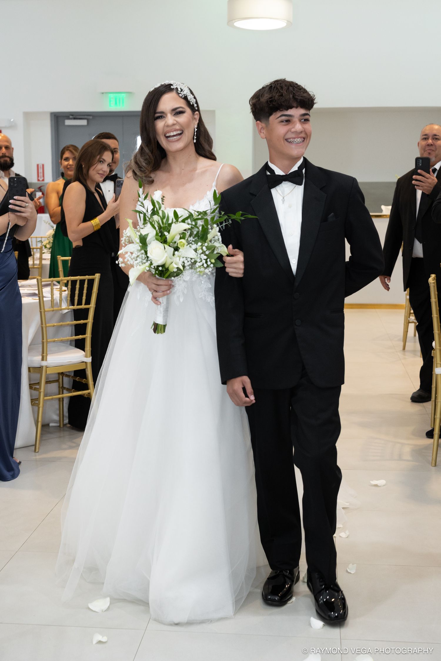 Bride in white ballgown holding bouquet walks down aisle with young man in black tuxedo at elegant wedding ceremony.