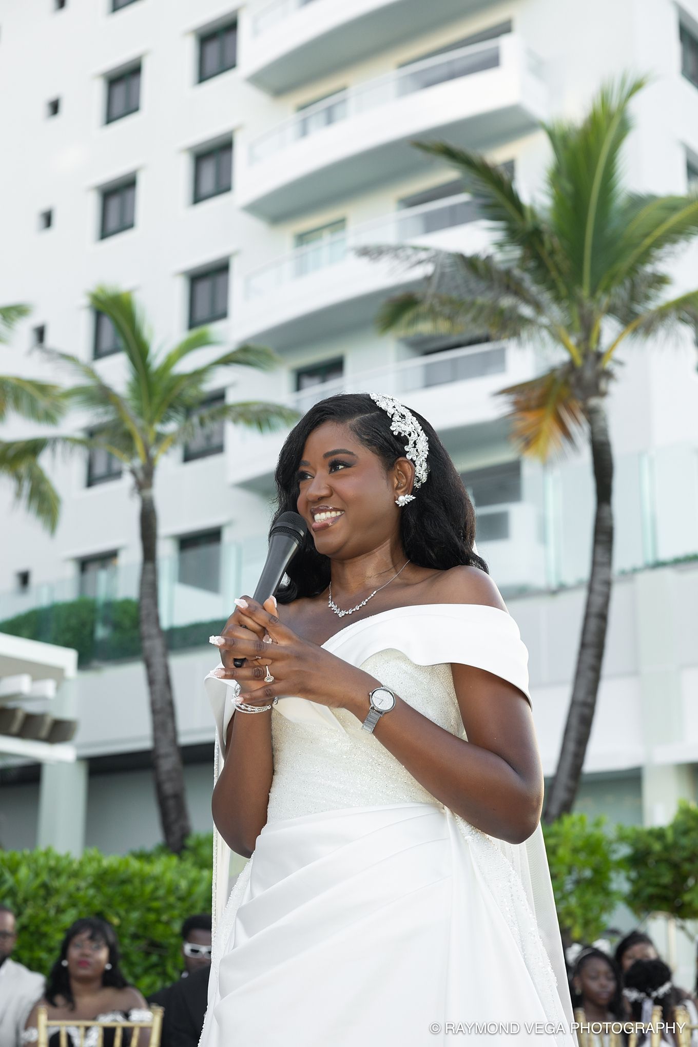 Smiling bride in white off-shoulder gown speaks at outdoor wedding ceremony near palm trees and hotel.