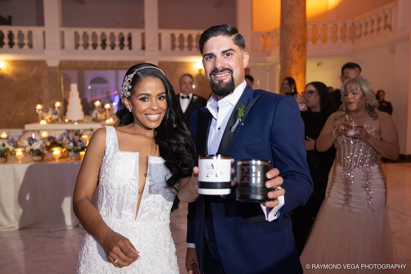 Bride and groom smiling at wedding reception, holding decorative candle, elegant ballroom venue with guests in background.