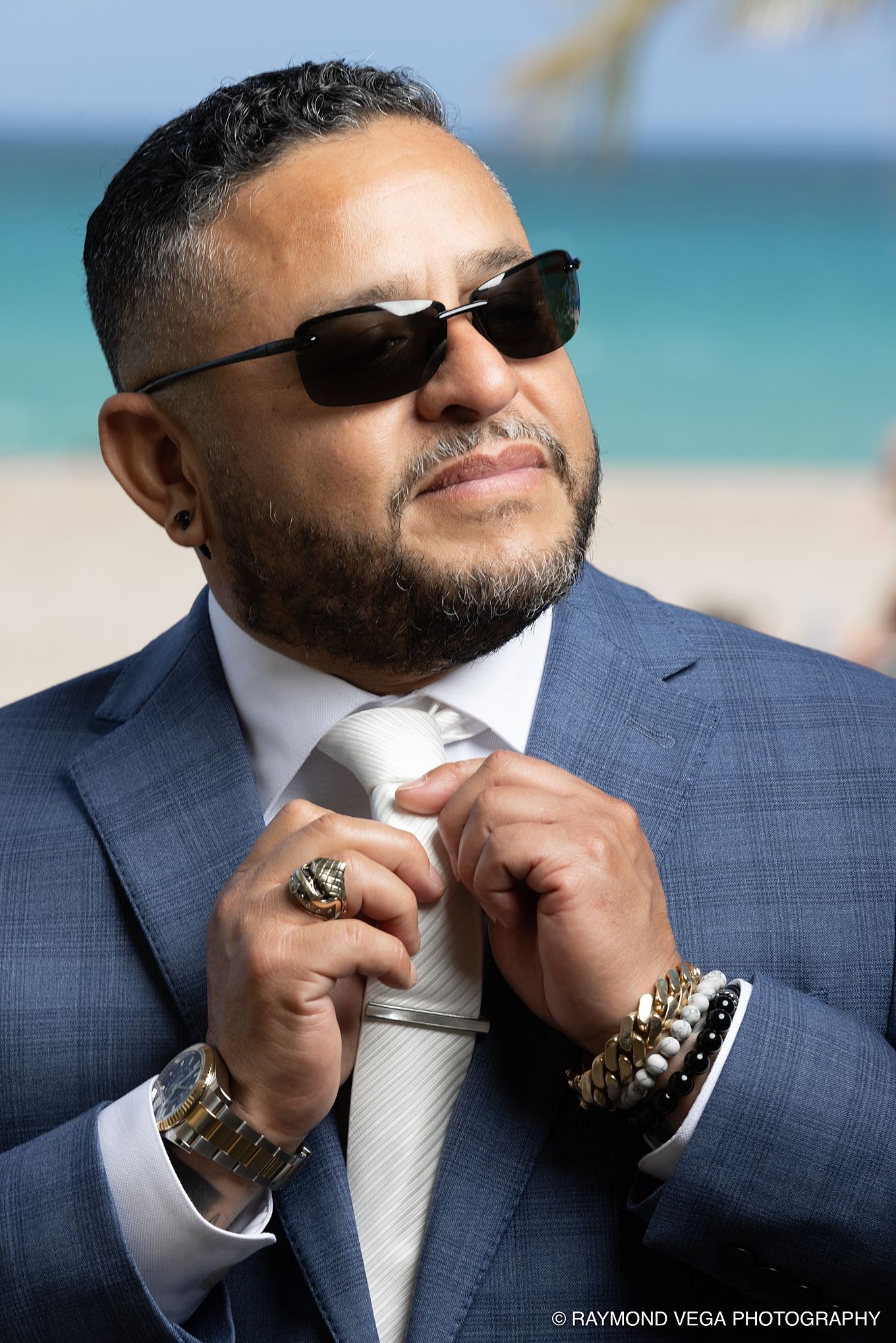 Man in blue suit adjusting tie on beach, wearing sunglasses, gold watch, and bracelets, photographed by Raymond Vega.