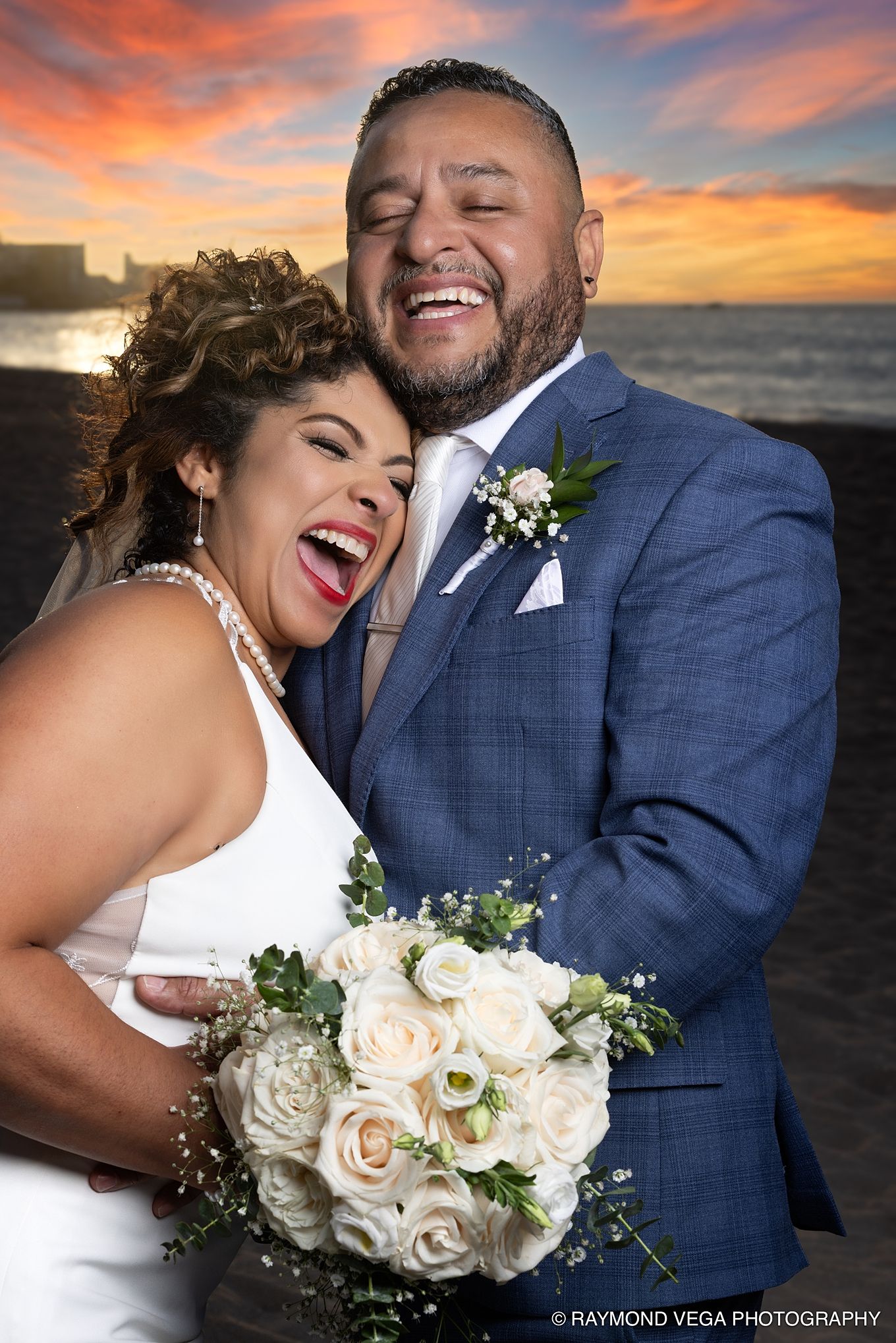 Joyful newlyweds laughing together at sunset, bride in white holding rose bouquet, groom in blue suit with boutonniere. at Courtyard