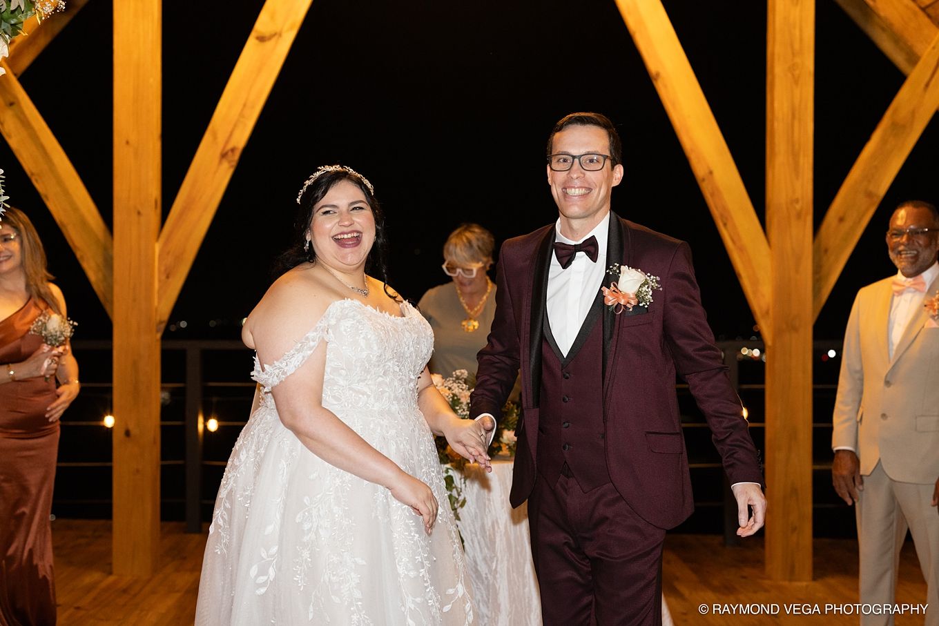 Bride and groom smiling joyfully during their wedding reception exit under wooden arch at night.