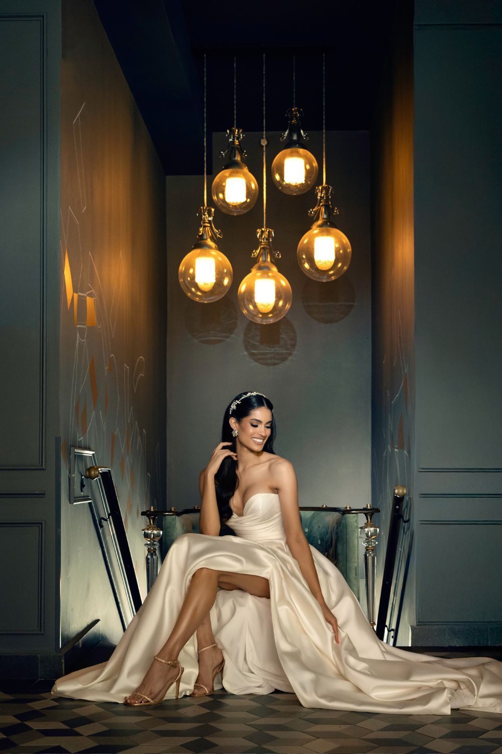 Elegant bride in a white satin gown seated under warm globe pendant lights in a luxurious, moody interior setting.