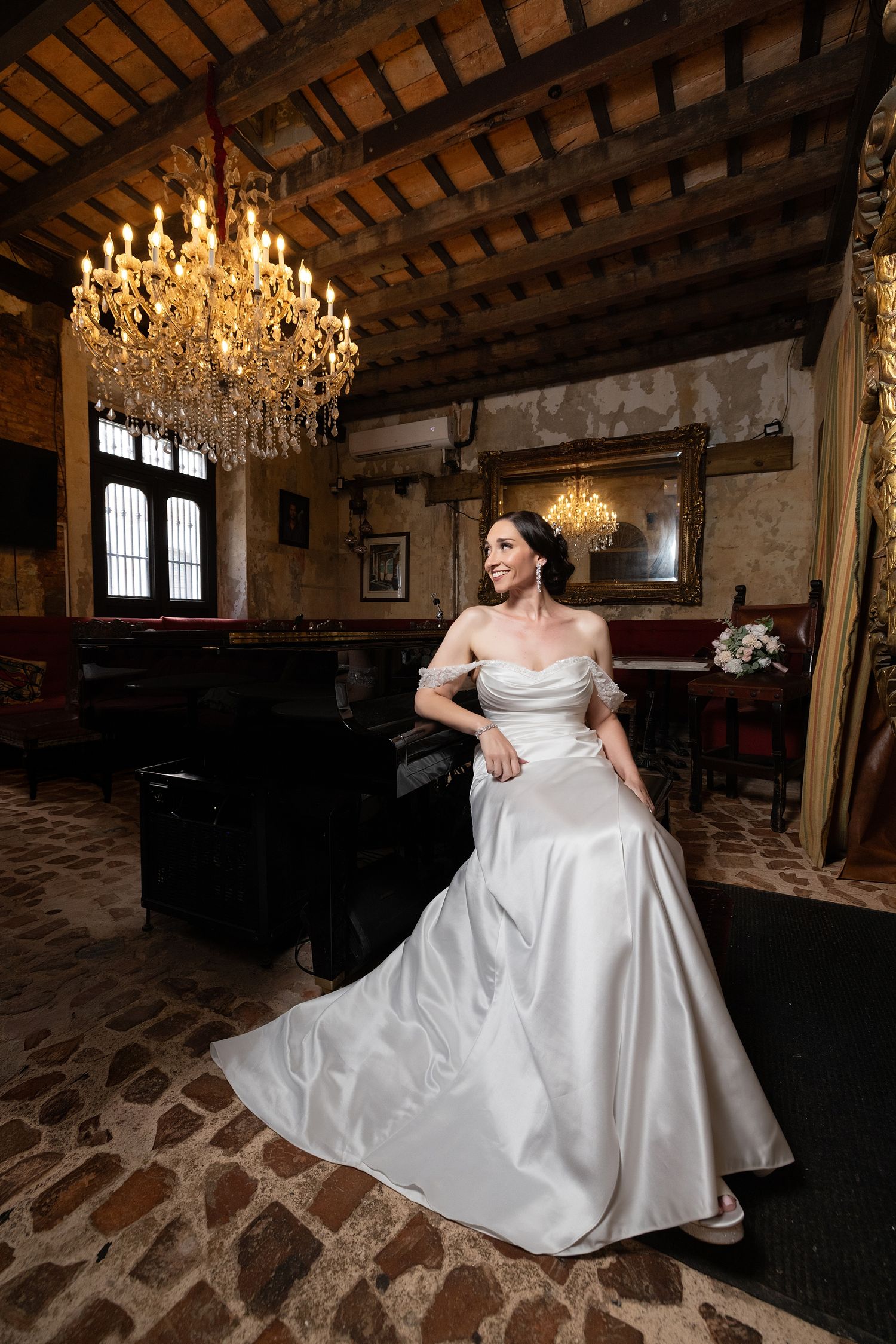 Bride in elegant off-shoulder satin gown poses in rustic venue with grand chandelier and stone floors.