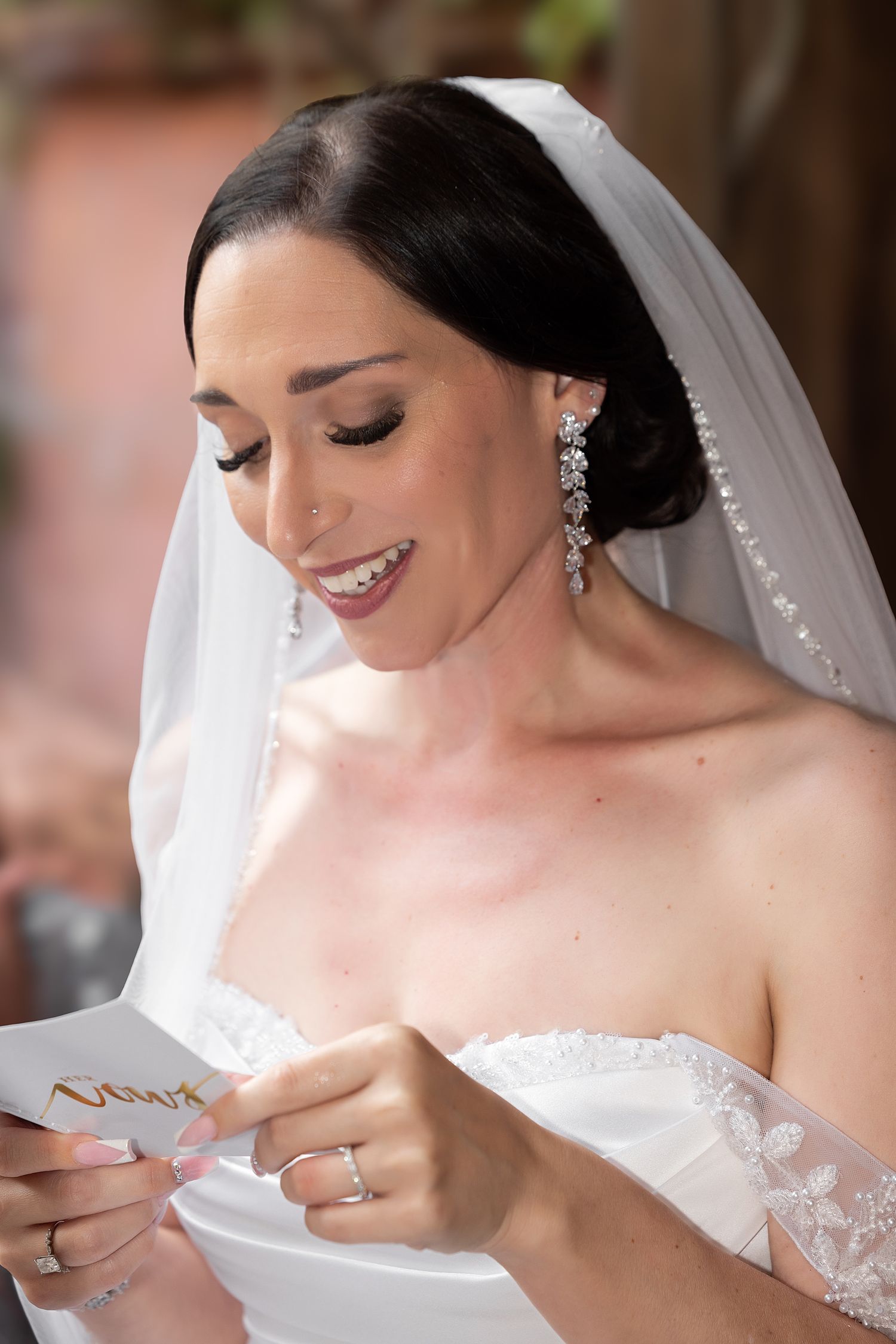 Smiling bride in white gown and veil reads a heartfelt letter, wearing elegant drop earrings on her wedding day.