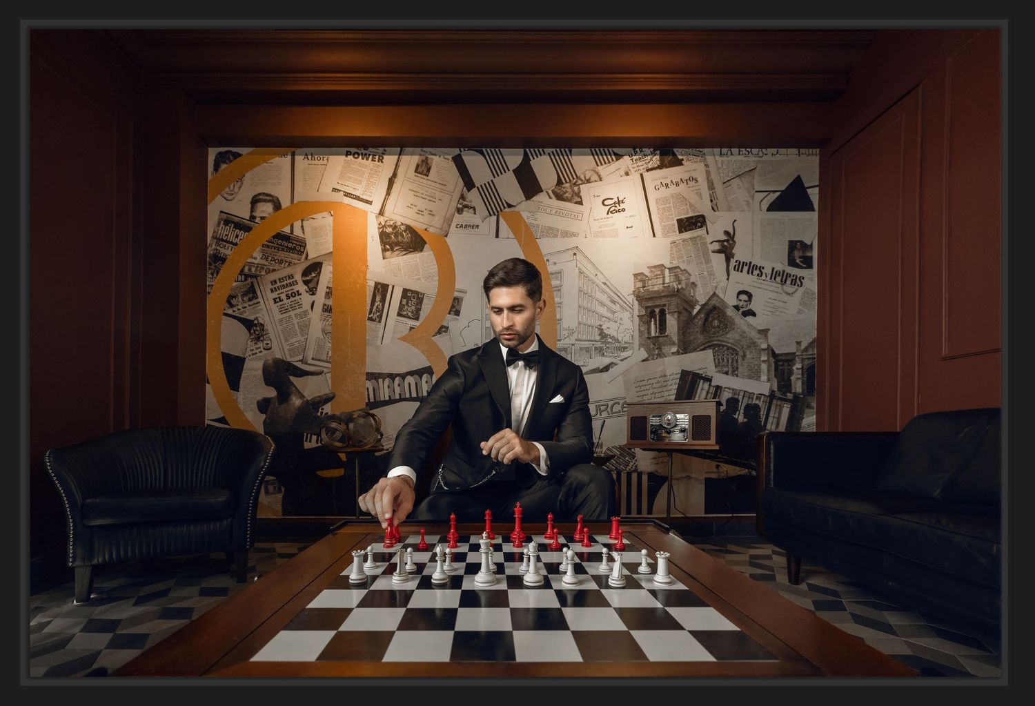 Man in tuxedo playing chess with red and white pieces in a sophisticated room with newspaper collage wall.