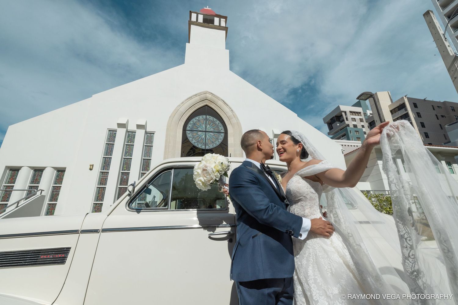 Bride and groom share a romantic moment outside a white church beside a classic white limousine on their wedding day.